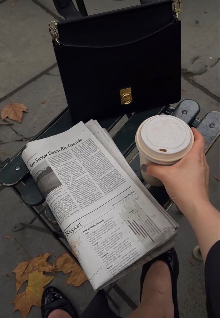 a person sitting on a bench reading a newspaper and holding a coffee cup in their hand