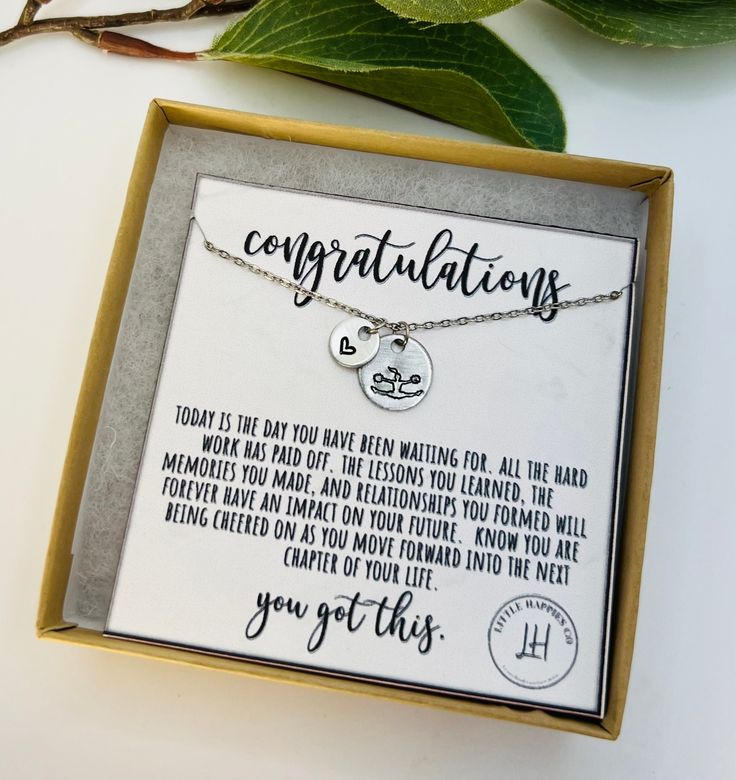 a necklace with congratulations written on it sitting in a box next to a plant and some leaves