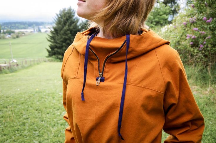 a woman in an orange hoodie is looking off into the distance with trees and grass behind her