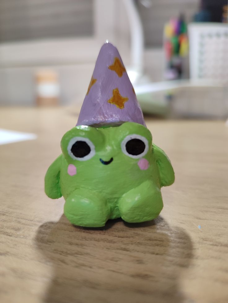 a green toy sitting on top of a wooden table wearing a purple hat with eyes