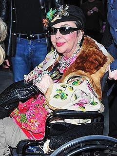 an older woman in a wheel chair wearing sunglasses and a fur coat, with other people behind her