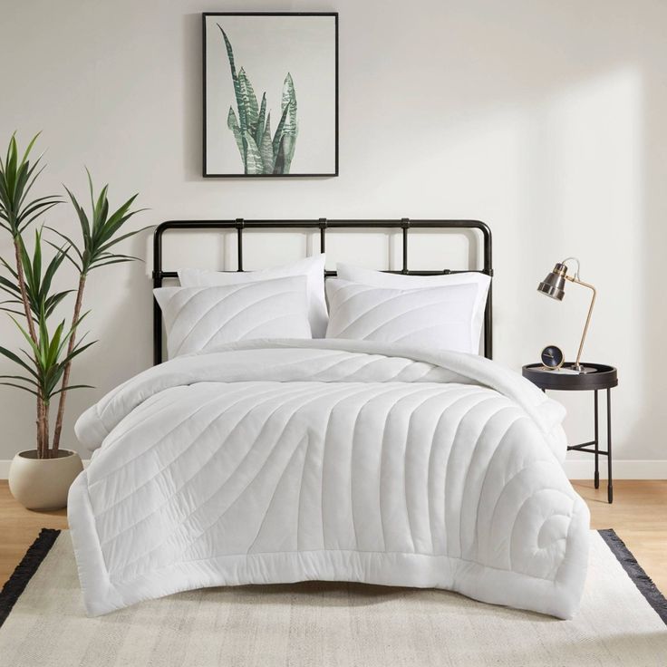 a bed with white comforter and pillows in a room next to a plant on the floor