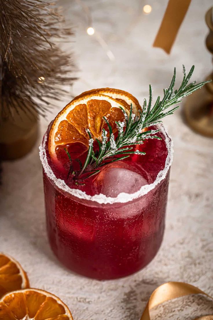 a red drink with an orange slice and rosemary garnish on the rim, surrounded by christmas decorations