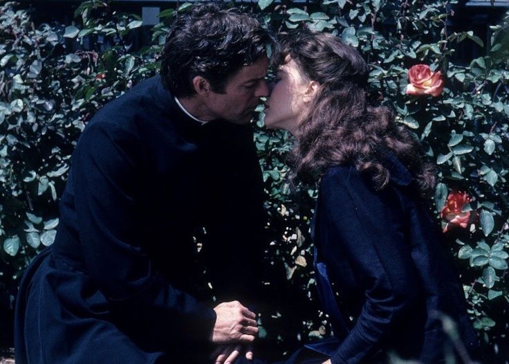 the man and woman are kissing in front of some bushes with red roses on it