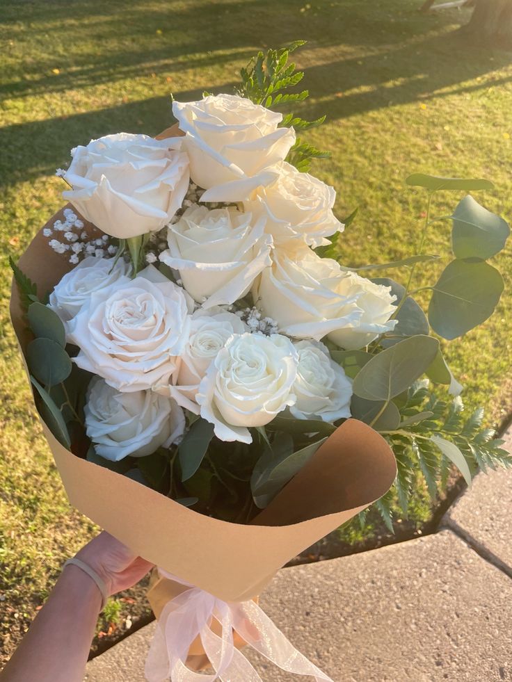 someone holding a bouquet of white roses on the sidewalk in front of some grass and trees