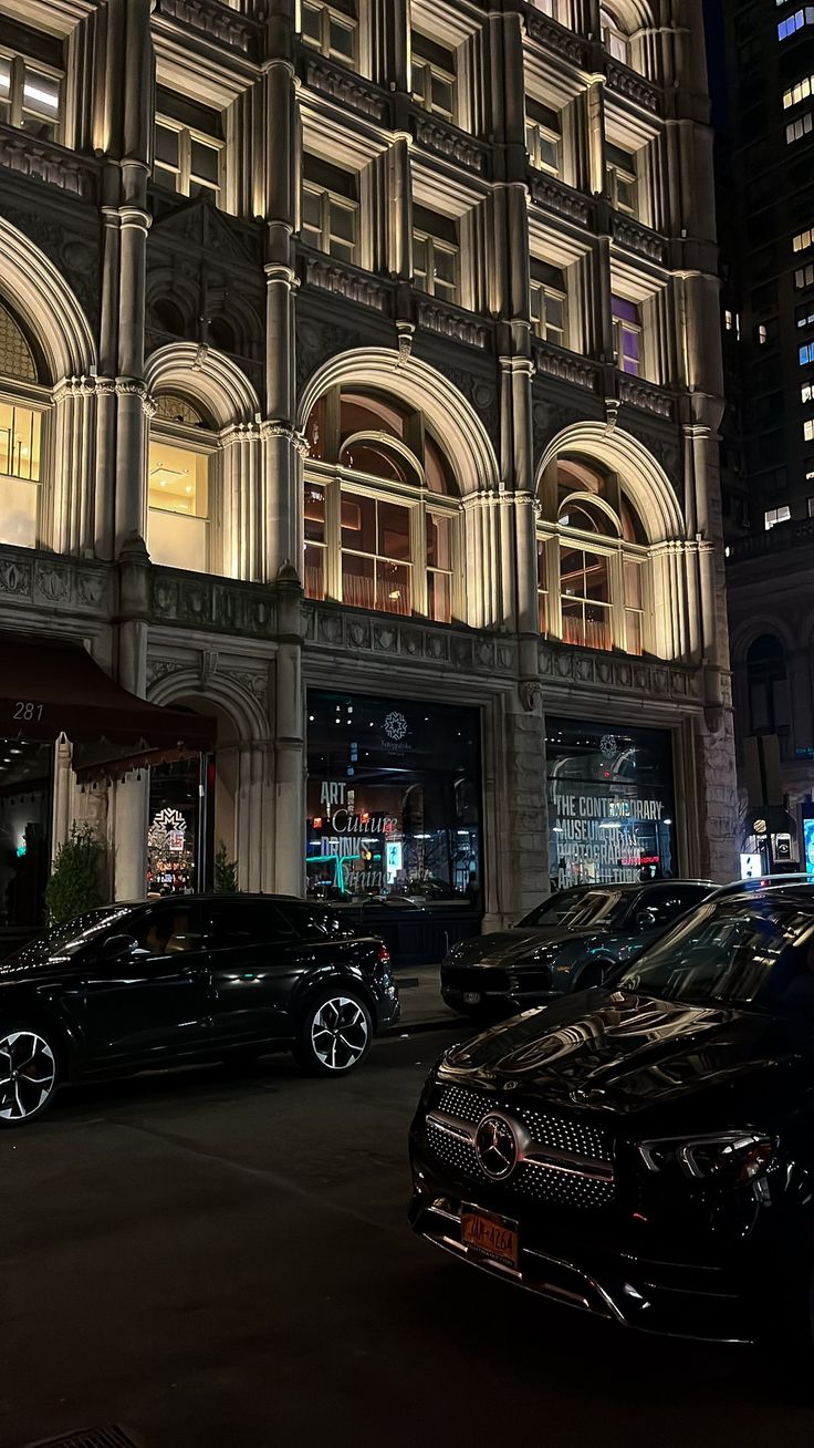 two black cars parked in front of a building at night with lights shining on the windows