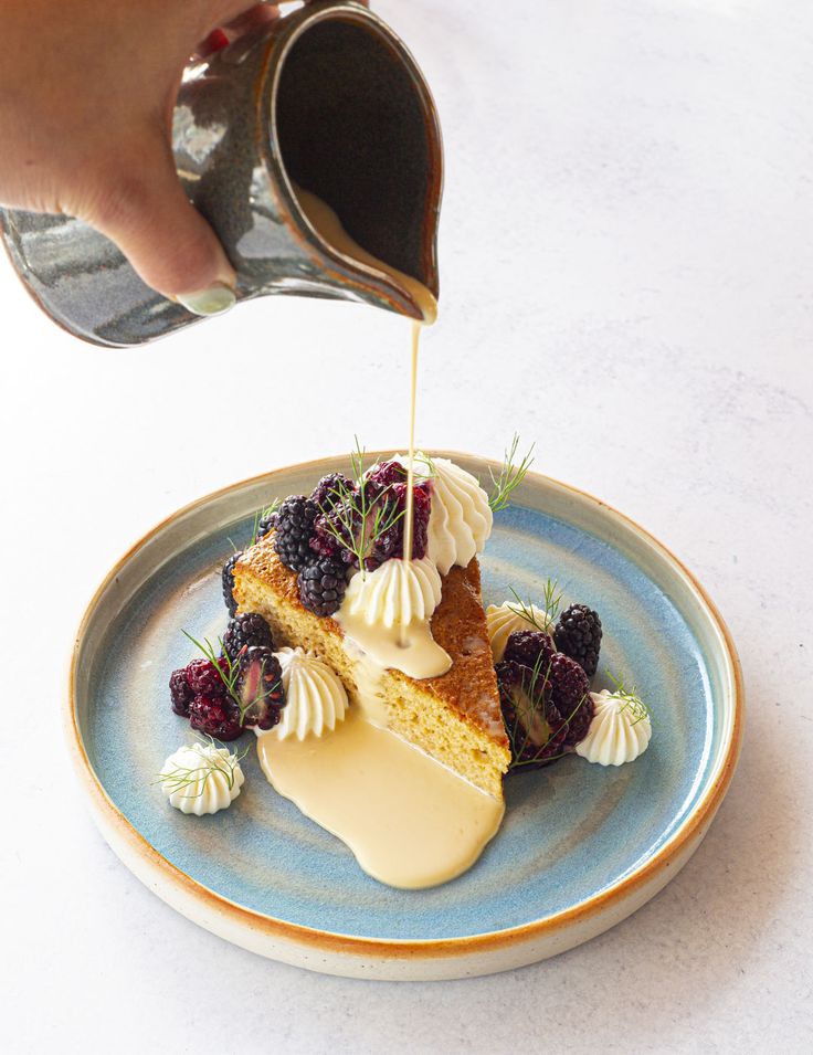 a person pouring syrup on a piece of cake with berries and whipped cream over it