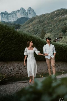 a man and woman holding hands while walking down a path with mountains in the background