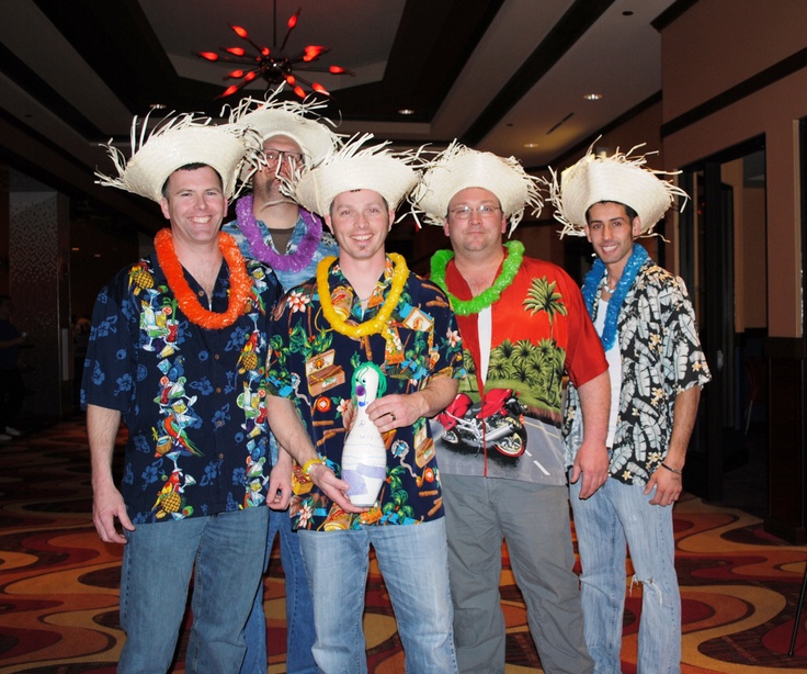 four men wearing hats and leis pose for a photo in front of a carpeted room