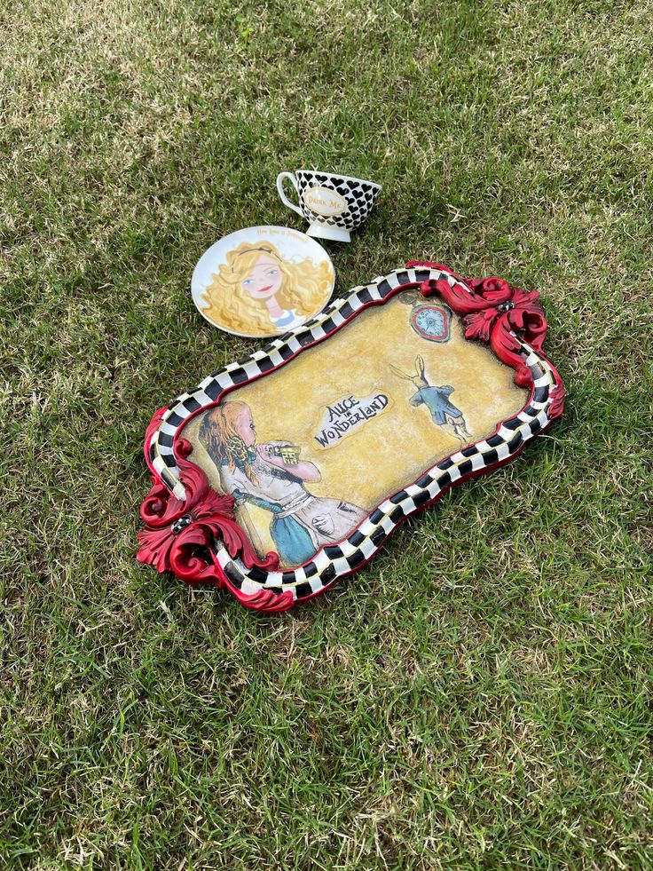 there is a plate and cup sitting on the grass next to a tray with an image of alice in it