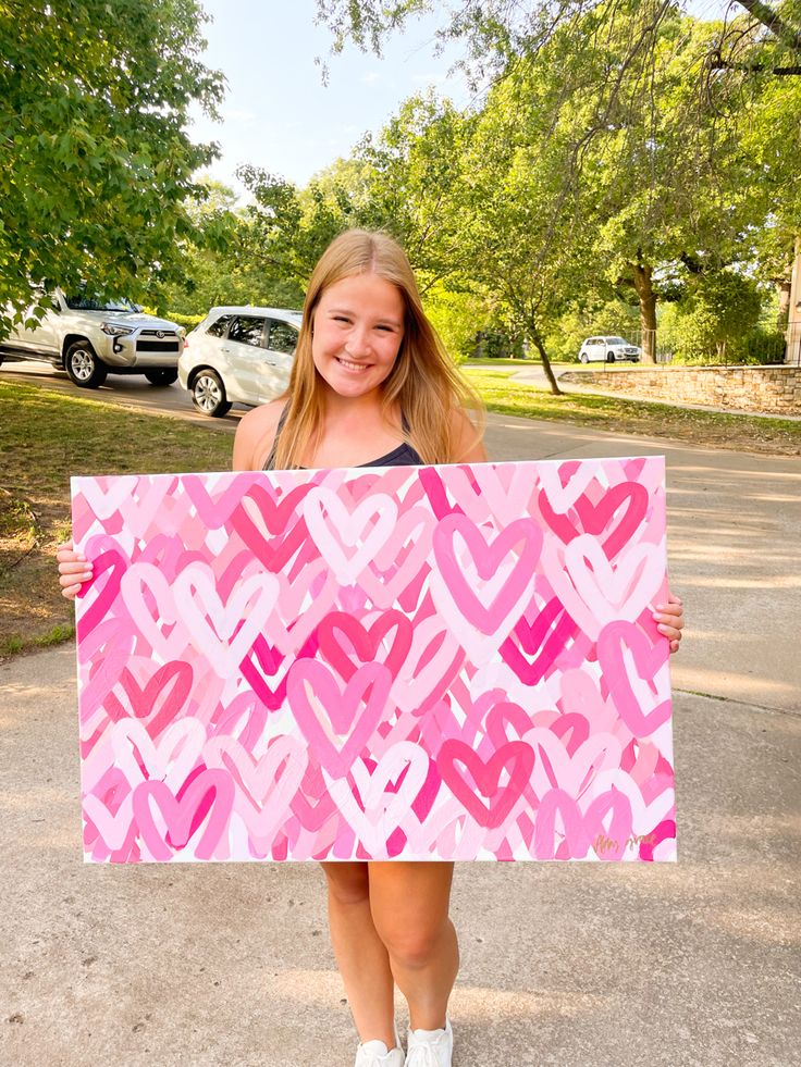 a girl holding up a pink and red painting with hearts on it in front of her