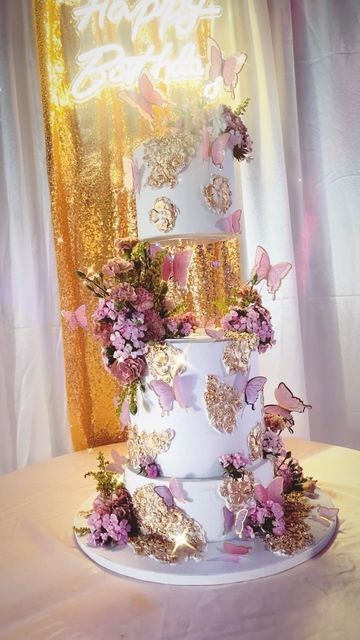 a three tiered white cake with pink flowers and butterflies on the top is surrounded by gold sequins