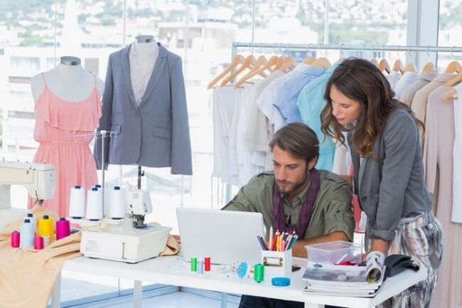 a woman and man working on a laptop computer in a clothing store with clothes hanging up behind them