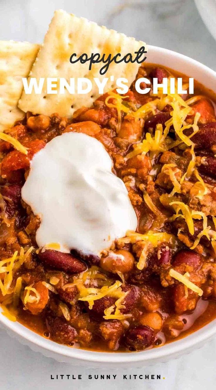 a white bowl filled with chili and topped with sour cream next to crackers on the side