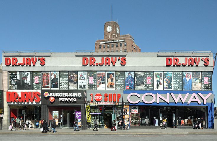 the front of a dr jay's store with people walking by it and a clock tower in the background