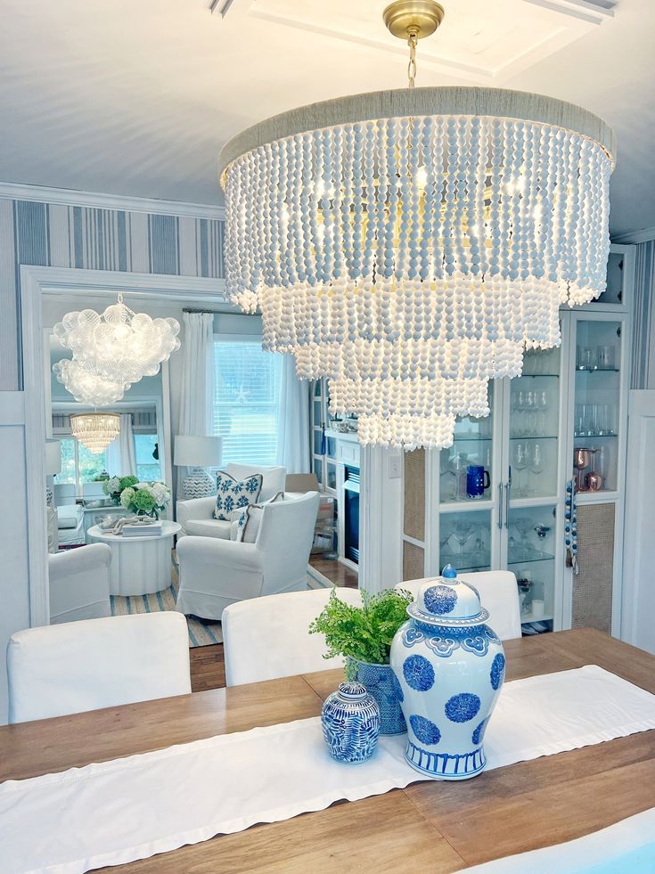 a chandelier hangs above a dining room table with white chairs and blue vases