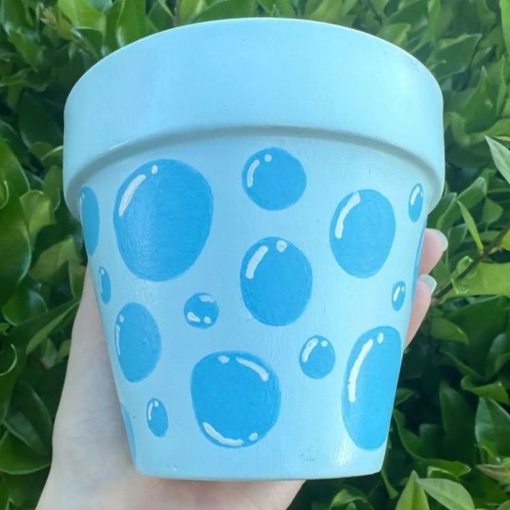 a hand holding up a blue cup with bubbles painted on the outside, and green bushes in the background