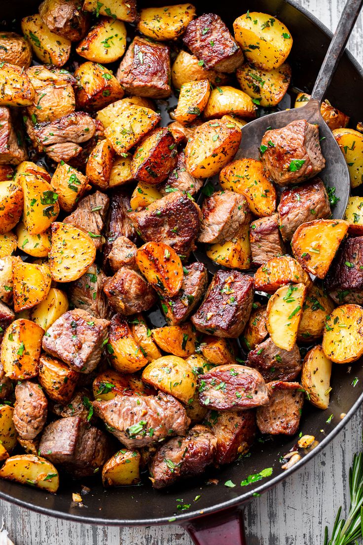 a skillet filled with potatoes and meat on top of a wooden table next to herbs