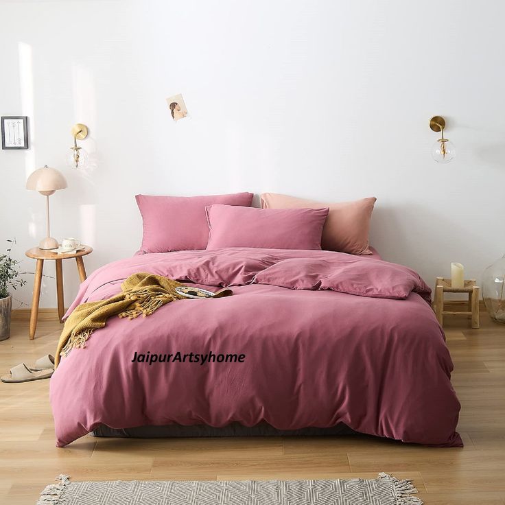 a bed with pink sheets and pillows in a white room next to a wooden floor