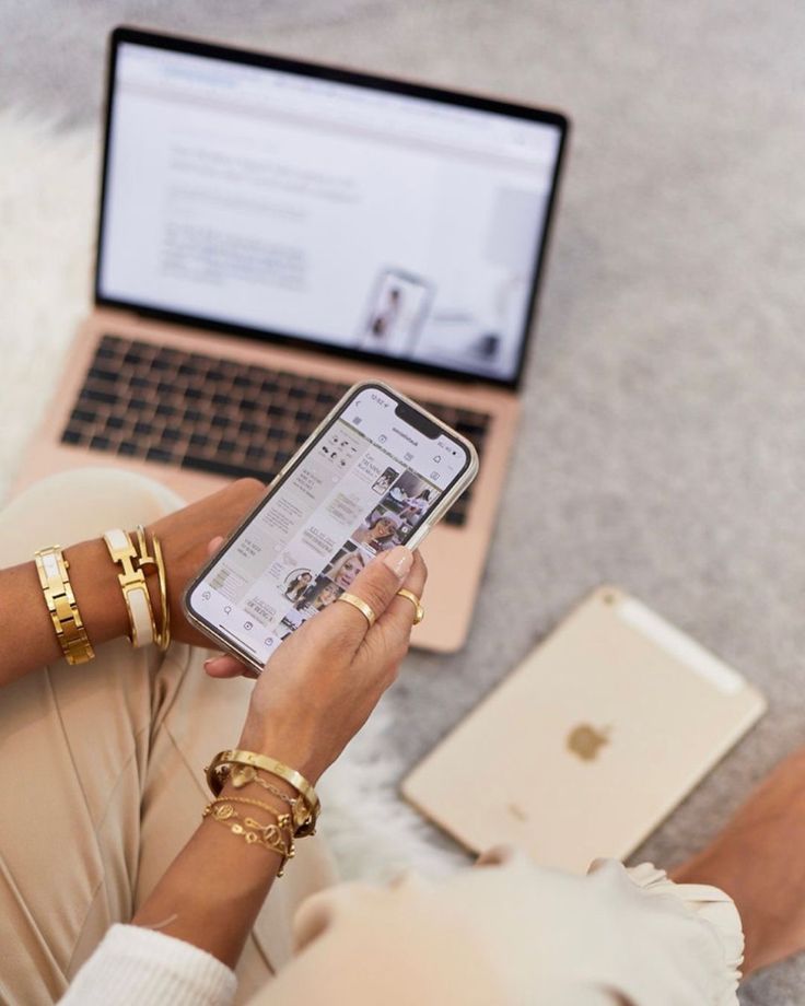 a woman sitting on the floor looking at her cell phone and laptop computer with gold bracelets