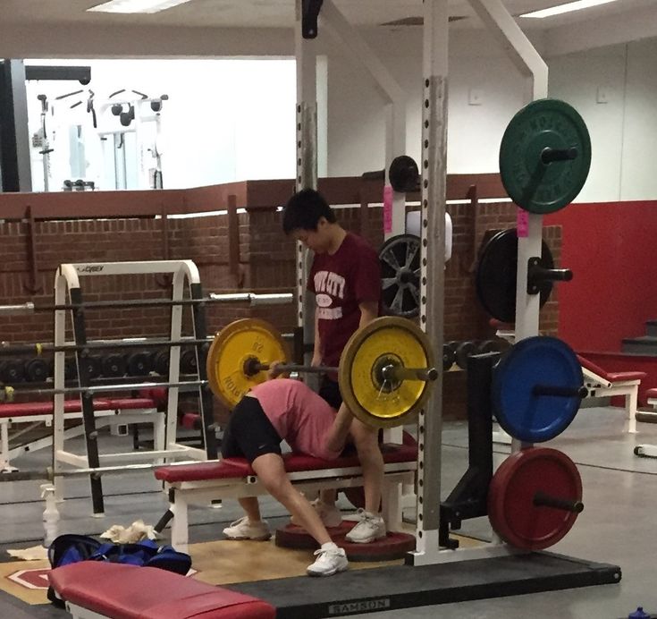 a man squats on a bench in a gym with another man working out behind him