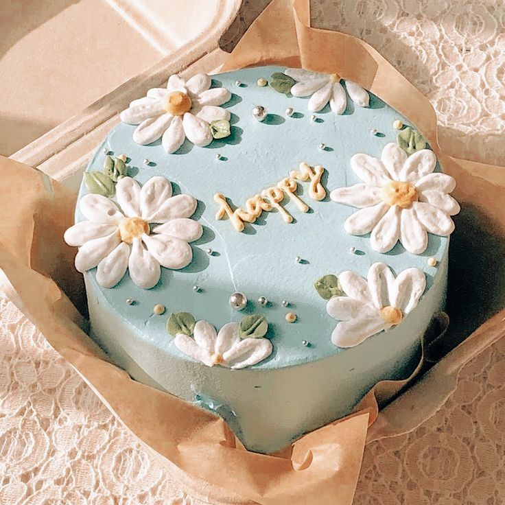 there is a blue cake with white flowers on it in a cardboard box next to lace doily