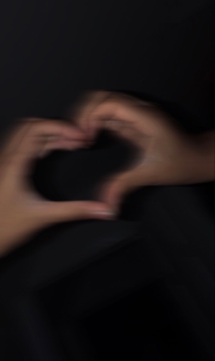 two hands making a heart shape with their fingers in the dark, against a black background