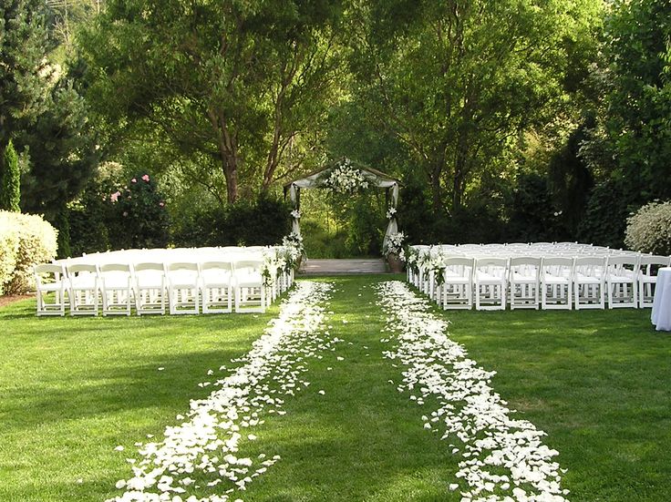 an outdoor ceremony setup with white chairs and flowers on the grass, surrounded by trees