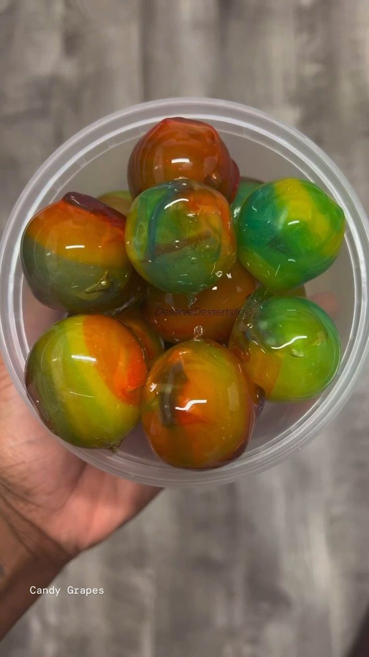 a person holding a plastic bowl filled with green and orange fruit on top of a wooden table