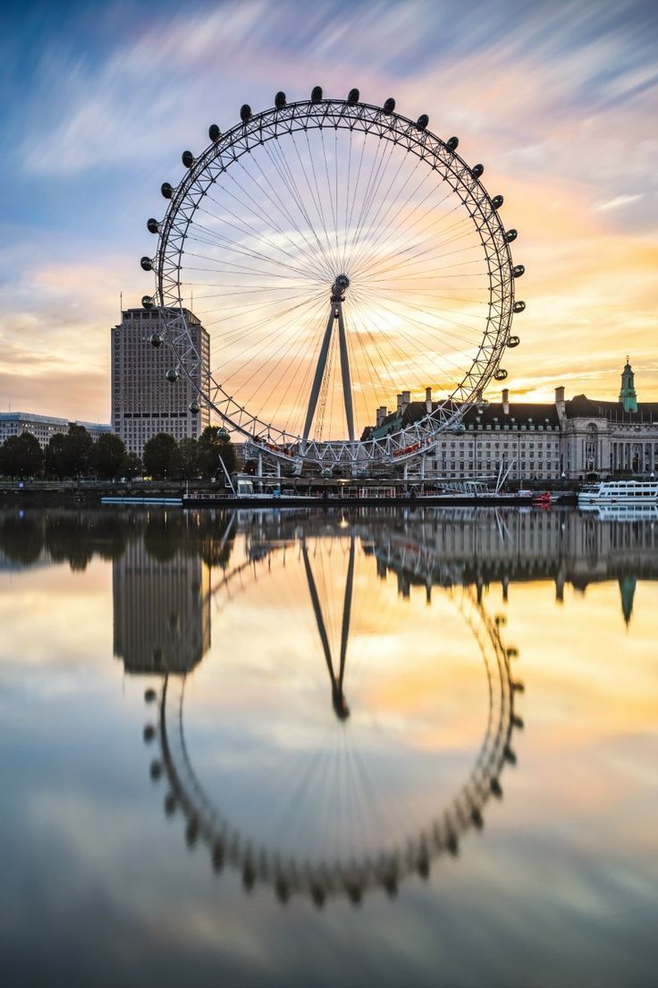 a ferris wheel is reflected in the water on a cloudy day with buildings behind it