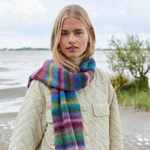 a woman standing on the beach wearing a multicolored knitted scarf and jacket