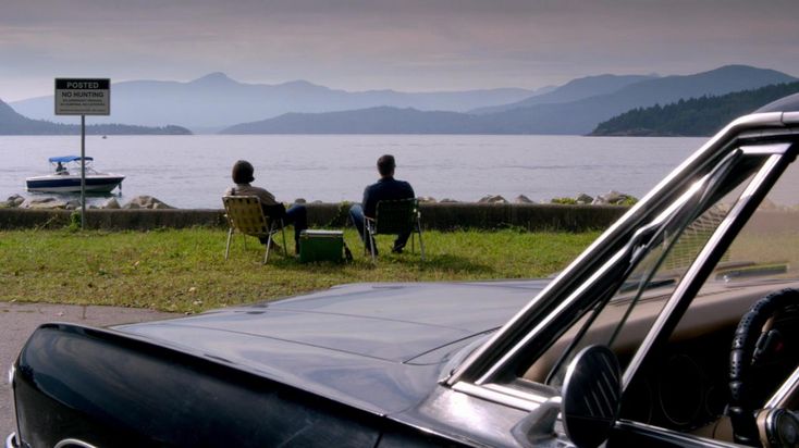 two people sitting in chairs on the side of a road next to a body of water