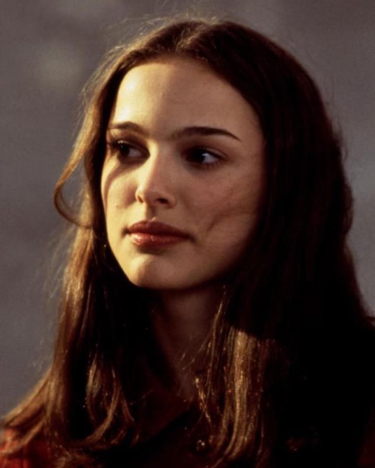 a woman with long brown hair and blue eyes looks off into the distance while standing in front of a wall