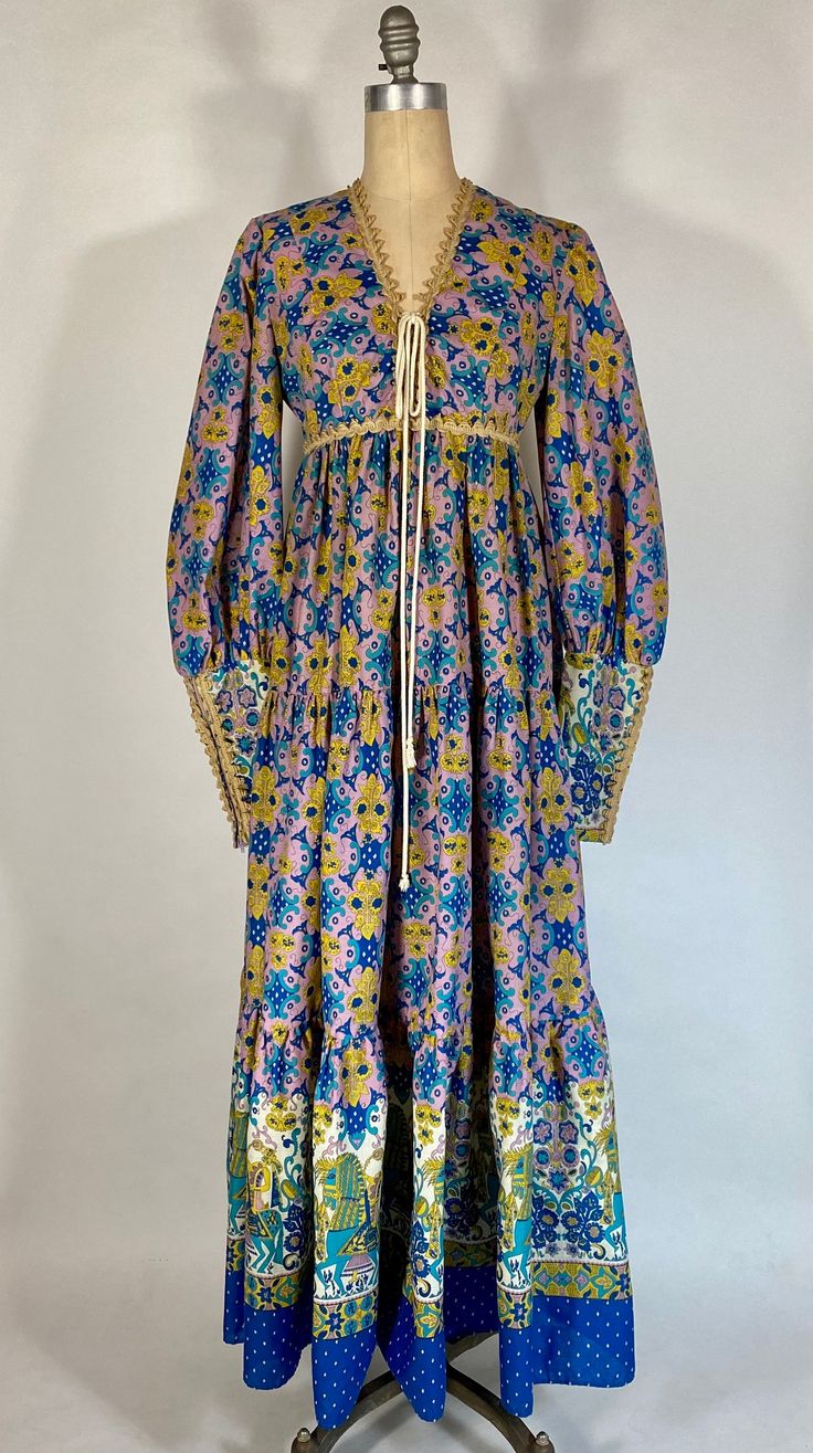 This early Gunne Sax dress is sure to turn heads! Whether you collect Gunne Sax dresses, or not, this early version is surely spectacular. It has a strong Bohemian vibe, and would've blended in perfectly, in Haight-Ashbury, circa 1969. It is made from a lightweight cotton or cotton blend fabric that features a vivid and busy print in blues, lilac, yellow and white. The 'vibe' of the pattern is very Persian and I believe it is officially called, or referred to as 'The Horseman Print' because it features a fanciful knight or horseman, atop his steed, and holding a feather in his hand. The cut and silhouette are somewhat Renaissance-inspired, with an empire style high waistline featuring a lace-up lower neckline with cotton cord lacing in ivory. The sleeves are incredible, puffed down to mid- Vintage Floral Print Maxi Dress For Evening, 1970s Style Maxi Dress With Vintage Print, 1970s Maxi Dress With Vintage Print, Retro Long Sleeve Festival Dresses, Retro Long Sleeve Dresses For Festival, Fitted Silk Festival Dress, Fitted Silk Dress For Festivals, Vintage Silk Dresses For Festival, Vintage Long Dress With Floral Print