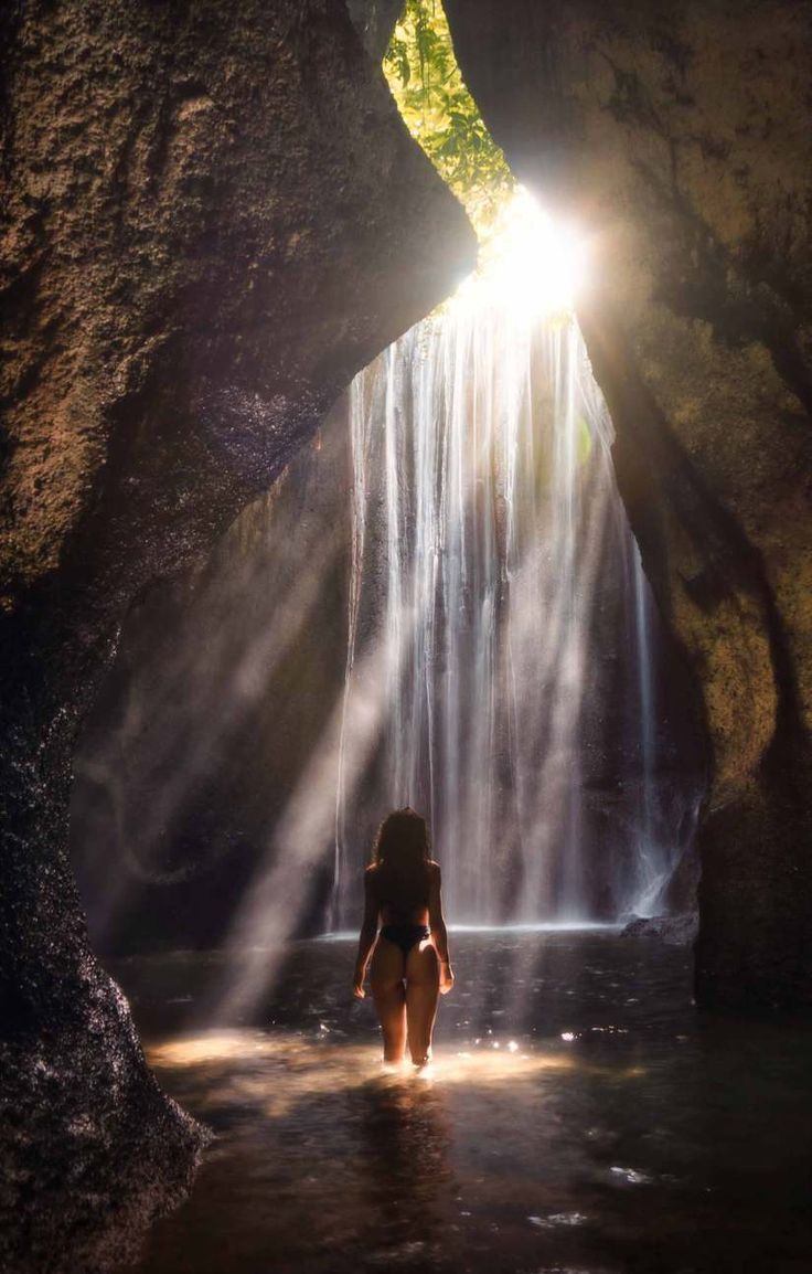 a woman standing in front of a waterfall with sunlight coming through the cave and her back turned to the camera