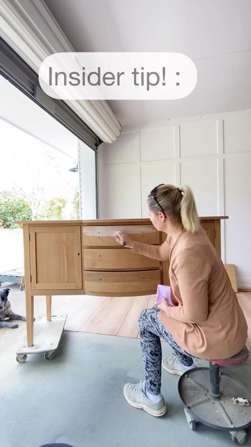 a woman sitting on top of a stool in front of a wooden dresser next to a window