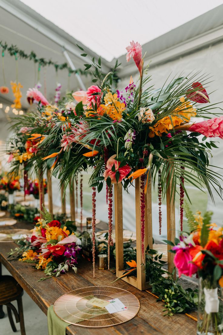 an arrangement of flowers and greenery is displayed on a long table in a greenhouse