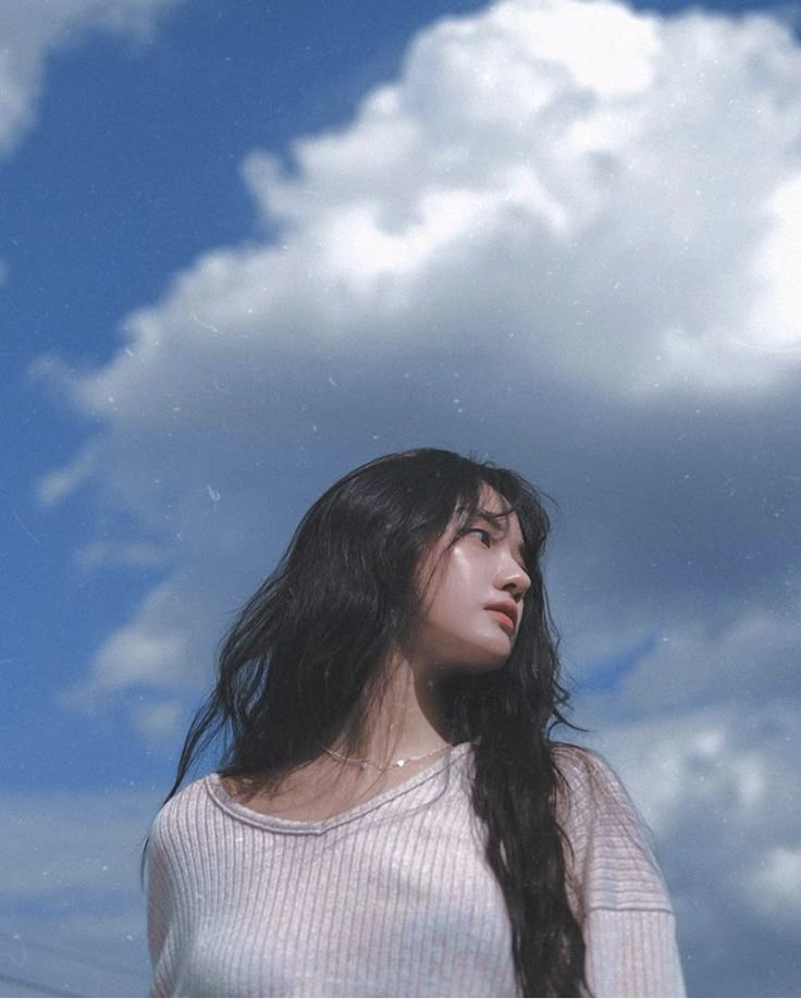 a woman with long hair standing in front of a blue sky and white fluffy clouds