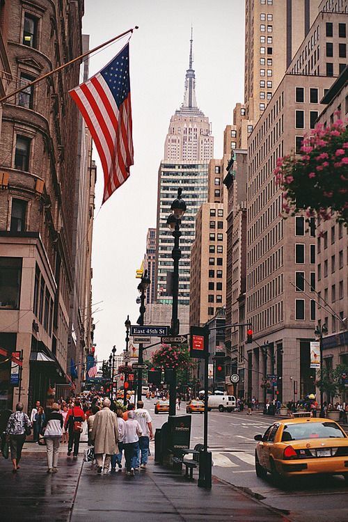 people walking down the street in front of tall buildings with an american flag on it