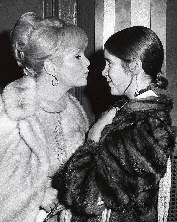 two women standing next to each other in front of a wooden door wearing fur coats