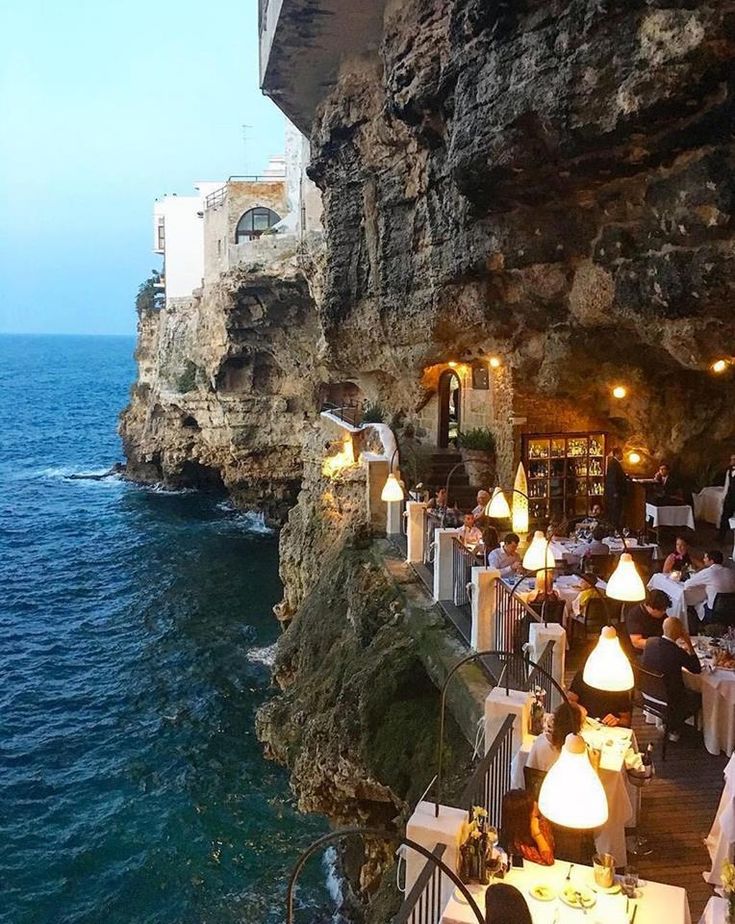 people are sitting at tables on the cliff by the ocean with lights hanging from them