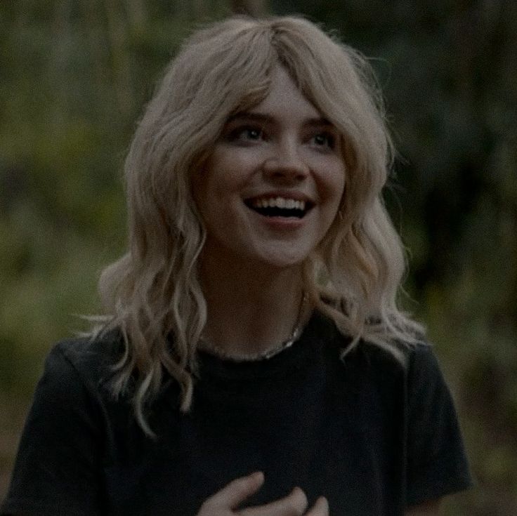 a woman with blonde hair smiling and wearing a black t - shirt in the woods