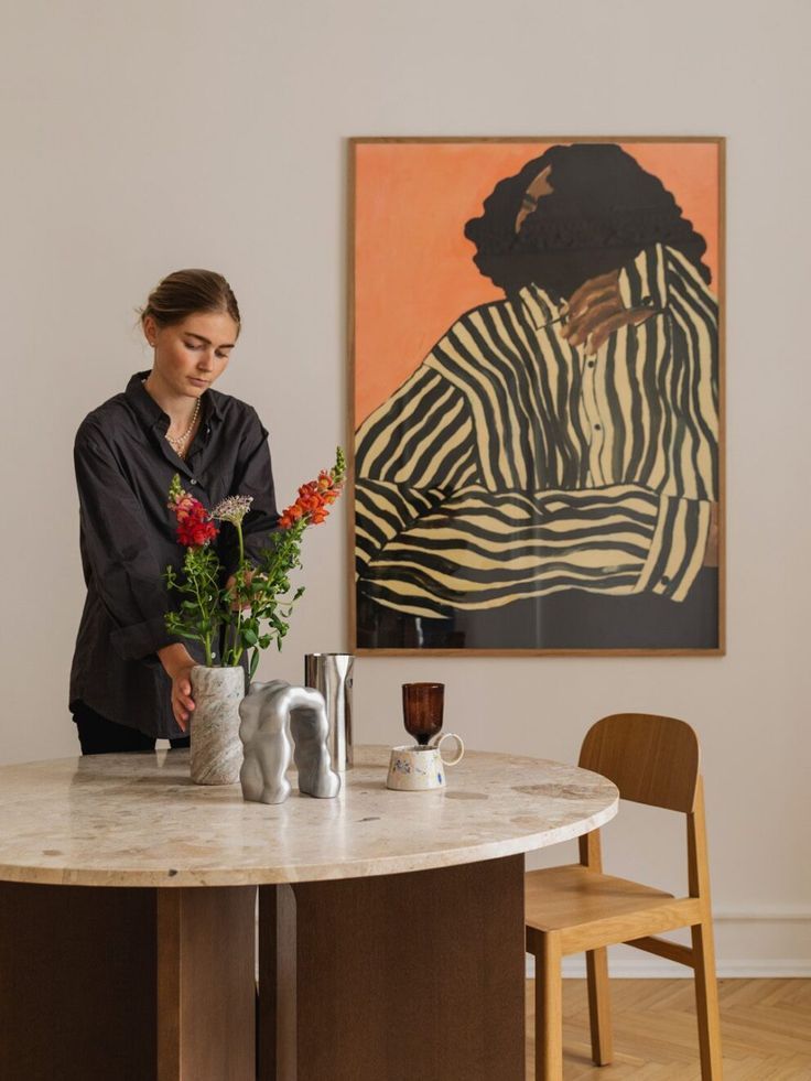 a woman standing at a table with vases and flowers in front of a painting