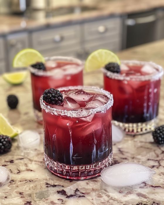 two glasses filled with blackberry margaritas on top of a marble counter next to lemon wedges