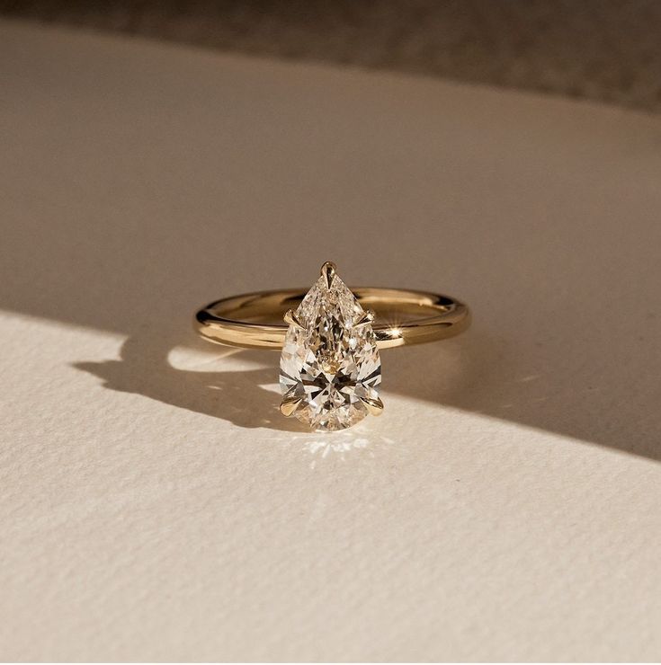 a yellow gold engagement ring with a pear shaped diamond on the side, sitting on a white surface