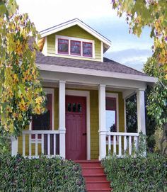 a small yellow house with red door and white railings on the front porch, surrounded by greenery