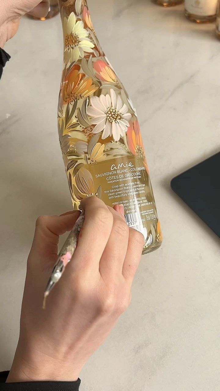 a person is holding a bottle with flowers on it and writing on the label in front of them