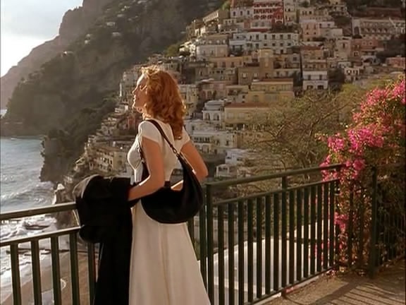 a woman in a white dress standing on a balcony next to the ocean and buildings