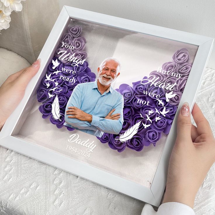 a person holding a photo in a box with purple paper flowers on the bottom and an image of elderly man