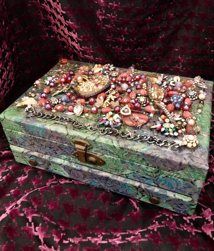 an ornate box is covered with beads and other jewelry on a pink velvet cloth background